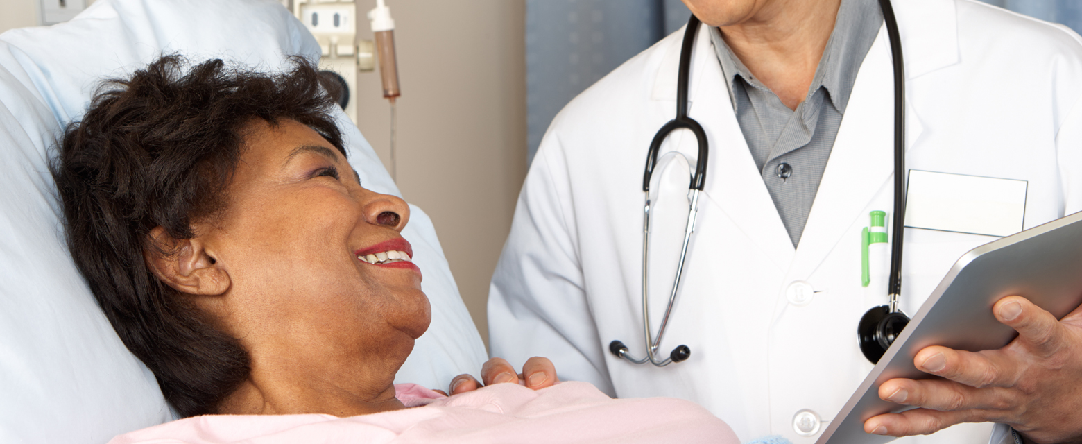 An adult African American female laying down in a hospital bed and smiling at her doctor who has his hand placed on her shoulder