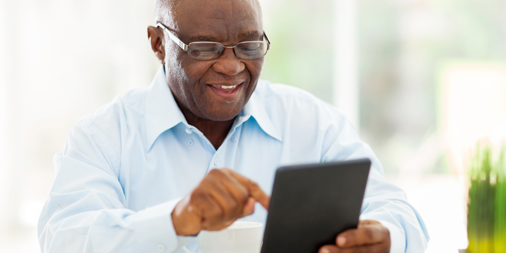 An adult African American male wearing a long sleeved collared shirt and glasses using a tablet and smiling